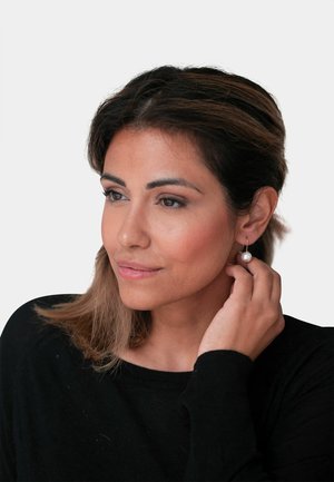 Goldene Creolen mit einer hängenden Perle, getragen von einer Frau mit mittelbraunem Haar, die einen schwarzen Pullover trägt, vor einem weißen Hintergrund.
