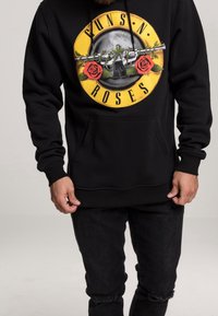 Homme portant un sweat à capuche noir Guns N' Roses avec un logo de pistolets croisés et des roses rouges sur un cercle jaune, associé à un jean noir déchiré.