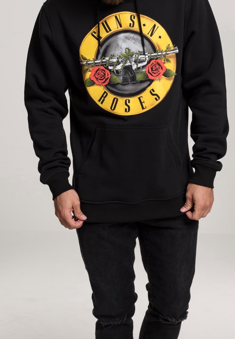 Homme portant un sweat à capuche noir Guns N' Roses avec un logo de pistolets croisés et des roses rouges sur un cercle jaune, associé à un jean noir déchiré.
