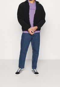 Svart kordjacka, lila T-shirt med text, blå jeans och svarta Vans-sneakers med vita detaljer. Hela kroppen syns, stående pose.