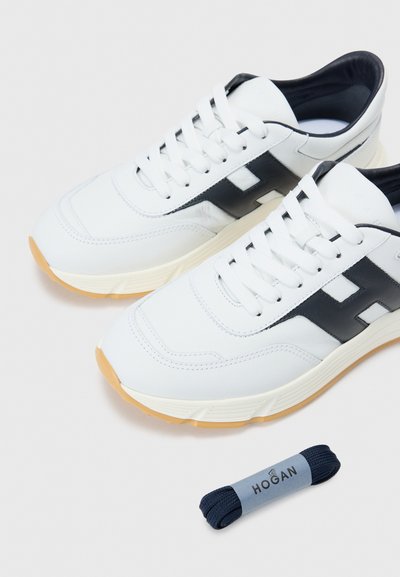 Baskets en cuir blanc avec des accents noirs, présentant des surfaces texturées et une semelle crème. Comprend un lacet de remplacement navy roulé.