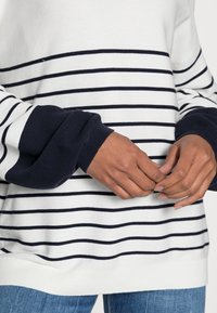 Person som bär en vit tröja med svarta horisontella ränder och marinblå ärmslut, med händerna löst knäppta över blå jeans.