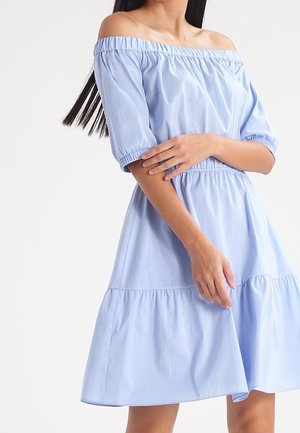 Vrouw draagt een lichtblauwe off-shoulder jurk met een elastische halslijn, korte pofmouwen en een geplooide, gelaagde rok.