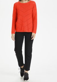 Rød strikket sweater med en chevronmønster, rund halsudskæring og lange ærmer, parret med sorte bukser og sorte sneakers.