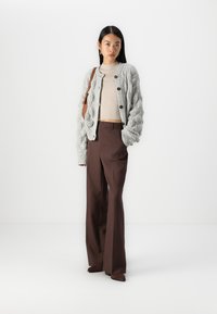 Cardigan gri tricotat cu un model texturat, pulover cu maneci lungi cu dungi dedesubt și pantaloni maro cu talie înaltă; modelul ține o geantă maro de umăr.