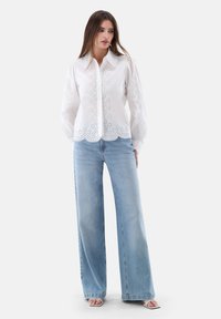 White long-sleeve blouse with eyelet detailing, scalloped hem, paired with light blue wide-leg jeans and white open-toe sandals.