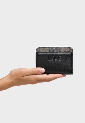 Hand holding a black leather TOUS wallet with a patterned beige and black top section against a light gray background.