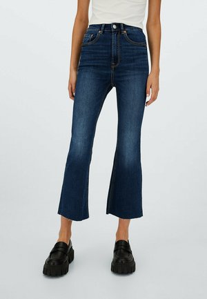 Femme portant un jean flare bleu foncé taille haute avec des ourlets bruts et des mocassins noirs épais, debout devant un fond uni.