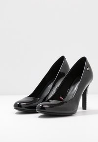 Pumps de charol negro de tacón alto con punta redondeada, plantilla acolchada y tacón transparente texturizado. Diseño minimalista con acento del logo.