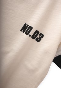 Kremskrāsas apģērbs ar melnu "NO. 03" izdruku. Mīksta tekstūra, ikdienišķs dizains ar apaļu kakla izgriezumu un īsām piedurknēm. Melni akcenti apakšmalā.