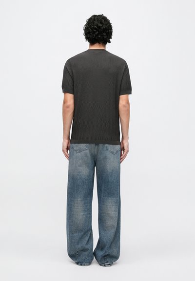 Sivá textúrovaná košeľa s krátkym rukávom v kombinácii s voľnými, vyblednutými modrými džínsami. Pohľad zozadu ukazuje jemný cikcak vzor na košeli.