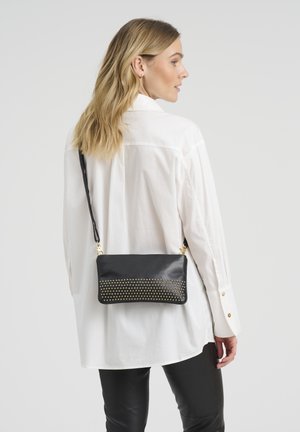 Žena sa plavom kosom dužine do ramena nosi preveliku bijelu košulju, crne hlače i crnu torbu s nitnama preko ramena.