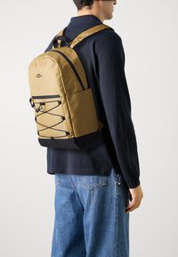 Zaino khaki realizzato in materiale resistente, con design a corda elastica, dettagli neri e spallacci imbottiti, indossato sulla schiena.