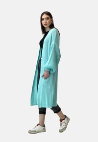 Cardigan lavorato a maglia turchese con maniche lunghe, apertura frontale e tasche laterali. Indossato sopra una maglia nera e abbinato a pantaloni neri e sneakers bianche.