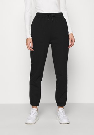 Vrouw draagt zwarte joggingsbroek met trekkoord, witte geribbelde longsleeve en witte sneakers, staand tegen een effen achtergrond.