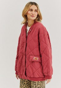 Prešívaná červená bunda so zapínaním na cvočky, rebrovanými manžetami a náprsníkou so zdobnou nášivkou. Nosená cez nohavice s leopardím vzorom.