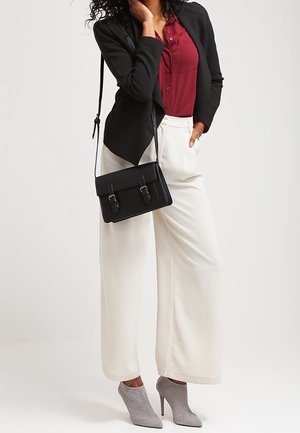Femme portant un blazer noir, un chemisier bordeaux, un pantalon blanc à jambes larges, des bottines grises à talons hauts et portant un sac noir en bandoulière.
