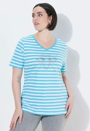 Femme aux cheveux courts et foncés portant un t-shirt rayé turquoise et blanc à col en V avec des visages de chiens et le texte « SELFIE », associé à un pantalon gris.