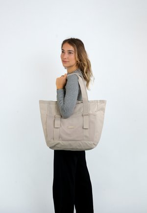 Beige Tragetasche aus strapazierfähigem Stoff, mit zwei langen Griffen, einer strukturierten Oberfläche und einem dezenten Markenschild auf der Vorderseite.