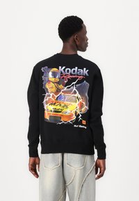 Sort sweatshirt med et farverigt racerdesign på ryggen, der fremhæver en kører og en bil, med teksten "Kodak Racing". Fremstillet af bomuldsstof.