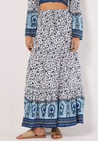 Jupe maxi blanche avec des motifs floraux bleus et des accents cachemire, dotée d'un design à niveaux et d'une taille élastique, assortie à des sandales beiges.