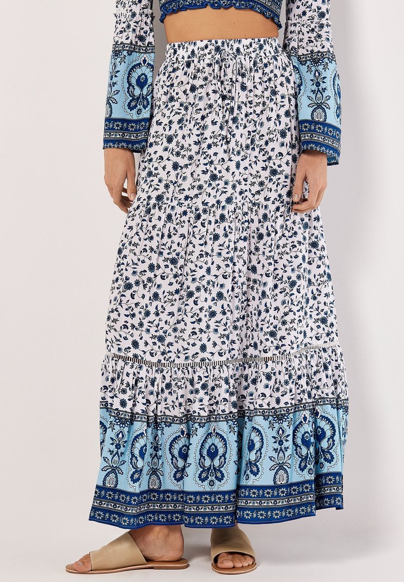 Jupe maxi blanche avec des motifs floraux bleus et des accents cachemire, dotée d'un design à niveaux et d'une taille élastique, assortie à des sandales beiges.