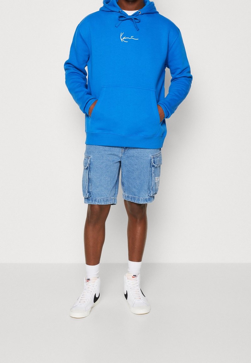 Homme portant un sweat à capuche bleu vif, un short cargo en denim clair, des chaussettes blanches et des baskets montantes blanches Nike avec un swoosh noir.