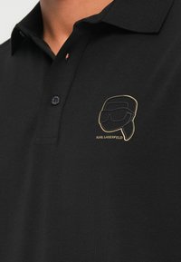 Čierne bavlnené polokošeľa s tri tlačidlami na zapínanie a vyšívaným zlatým logom na hrudi zobrazujúcim štýlizovanú tvár.