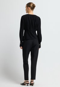 Completo nero su misura caratterizzato da una blusa plissettata e pantaloni slim-fit, con polsini bottonati e una silhouette strutturata. Accentuato con hardware dorato.