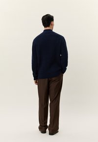 Moški stoji obrnjeni stran, oblečen v mornarsko modri pleteni pulover, rjave hlače in črne čevlje na svetli enobarvni podlagi.