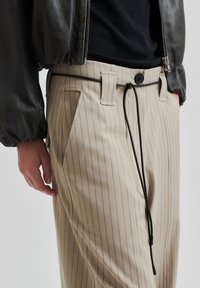 Beige, stripete bukser med en avslappet passform, med sort snor og knappelukking. Stoffet har en glatt tekstur.