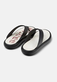 Mustat flip-flopit, joissa on kaksi leveää nauhaa, pehmustettu pohja ja valkoinen sisäpohja, jossa on punaista painettua tekstiä. Logo kohokuviona kantapäässä.