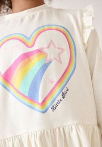 Roomkleurige top met een hartgrafiek met een regenboog en sterren. De stof is zacht en heeft ruches op de schouders.