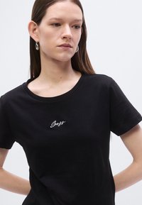 Črna bombažna majica z okroglim izrezom, na levi strani prsnega koša z belim vezenim napisom "Bess". Kratki rokavi, minimalističen dizajn.