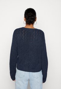 Pull en maille bleu marine avec un motif chevron texturé et des manches longues, doté d'un col rond et d'un ourlet côtelé. Associé à un jean bleu clair.