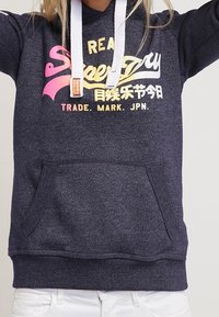 Sudadera azul marino hecha de tela texturizada, con un bolsillo tipo canguro, cordones blancos y estampados de logotipos multicolores en la parte delantera.