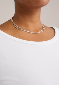 Detailní záběr osoby, která má na odhaleném krku dva tenké stříbrné náhrdelníky a bílé tričko s kulatým výstřihem.