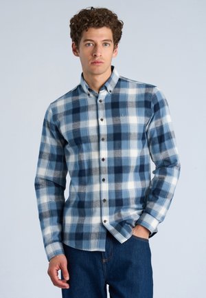 Ung mand iført en blå- og hvidternet skjorte med knapper og lange ærmer samt mørkeblå jeans, stående foran en lys baggrund.
