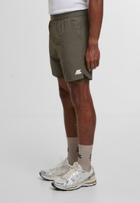 Pantalones cortos deportivos de color verde oliva con cintura elástica, con un logotipo blanco en el lateral, combinados con zapatillas gris claro y calcetines en tonos neutros.