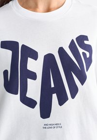 T-shirt blanc avec un grand texte bleu marine en gras qui dit "JEANS" avec un texte plus petit en dessous. Tissu doux et design classique à col rond.