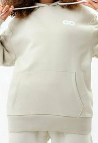 Personne portant un sweat à capuche beige avec une poche frontale, tenant les cordons près du cou, montrant un petit logo blanc sur la poitrine.