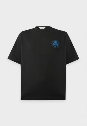 Homme portant un t-shirt noir avec un motif bleu sur la poitrine, un pantalon large foncé et des baskets beiges, debout devant un fond clair uni.