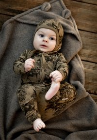 Brązowy kombinezon w wzory z dopasowanym kapeluszem, z motywami leśnymi. Miękka tkanina, dopasowany krój i wiązanie z przodu. Ułożony na szarym kocu.