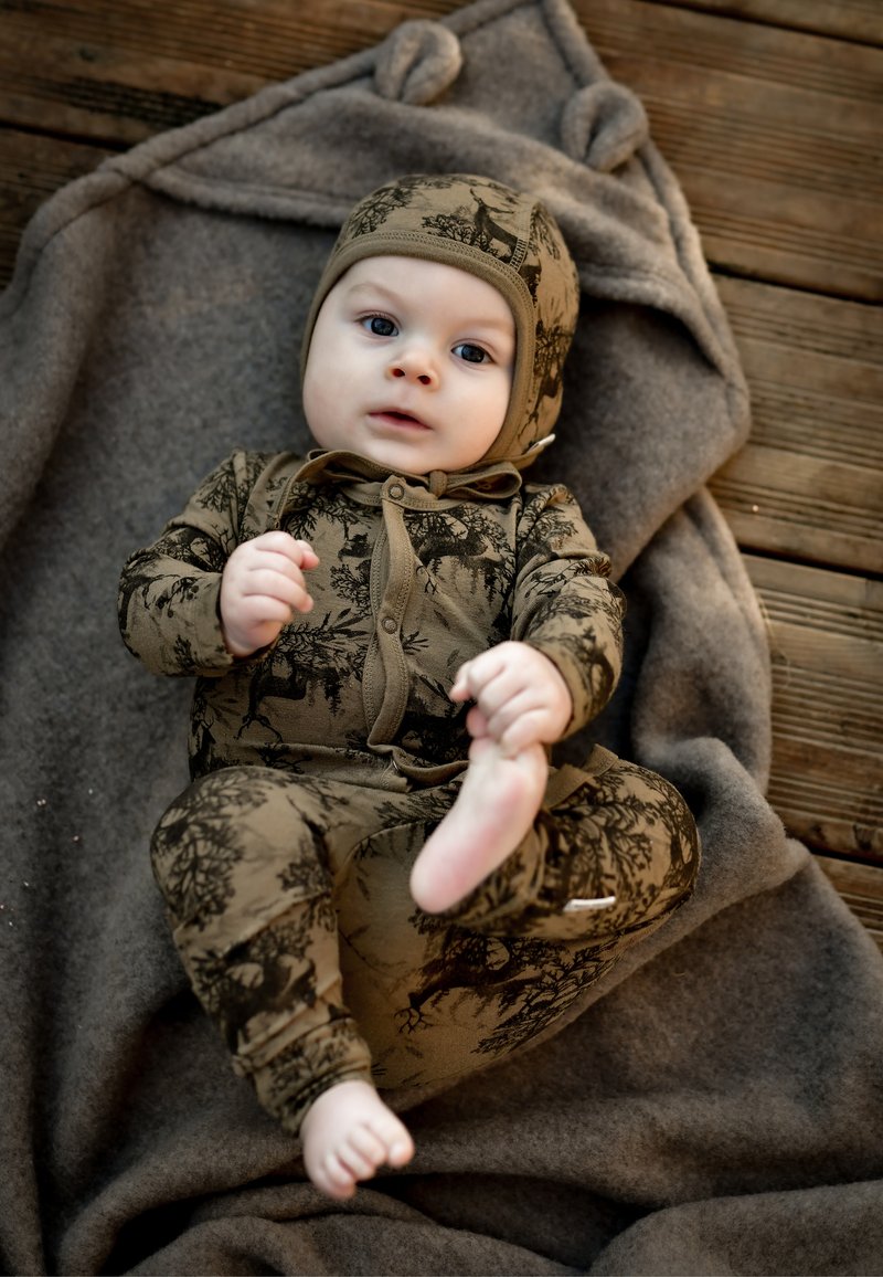 Brązowy kombinezon w wzory z dopasowanym kapeluszem, z motywami leśnymi. Miękka tkanina, dopasowany krój i wiązanie z przodu. Ułożony na szarym kocu.