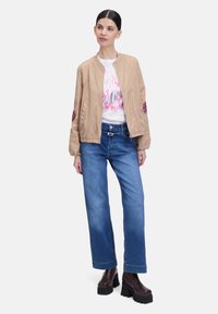 Beige bomberjack met geborduurde accenten, witte graphic t-shirt, blauwe wijde jeans en donkere platformlaarzen. Casual outfit.