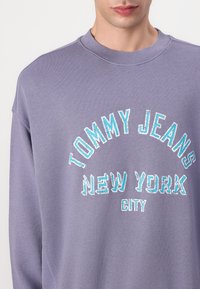 Grey sweatshirt featuring "TOMMY JEANS NEW YORK CITY" in blue and white embroidered lettering. Textured fabric with a round neck design.