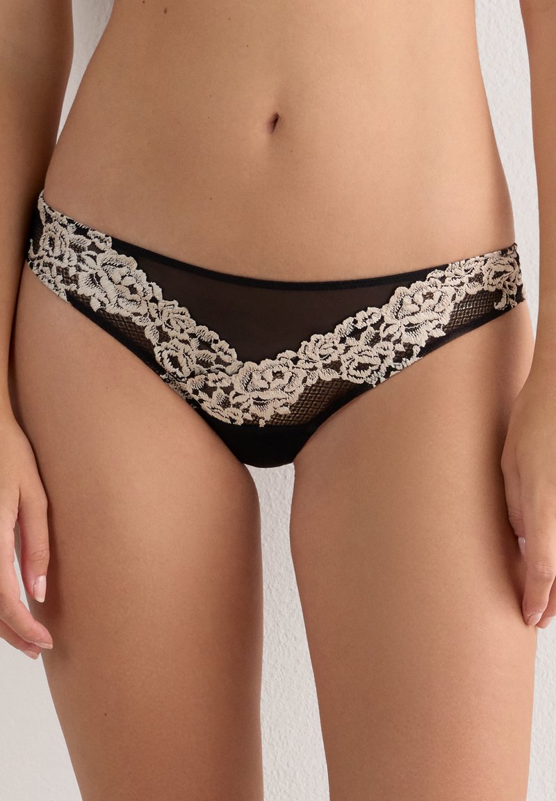 Sort bikini-stil undertøj med beige blomsterblonde detaljeret, båret af en person, der står mod en ensfarvet væg.