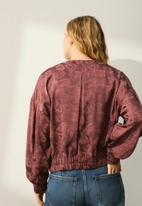 Chaqueta bomber burdeos con patrón botánico, cintura elástica y mangas oversize, combinada con vaqueros de mezclilla azul.