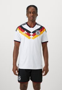 Mann trägt deutsches Fußballtrikot mit weißem Trikot, das schwarze, rote und gelbe Keile sowie schwarze Shorts mit Teamemblem zeigt.