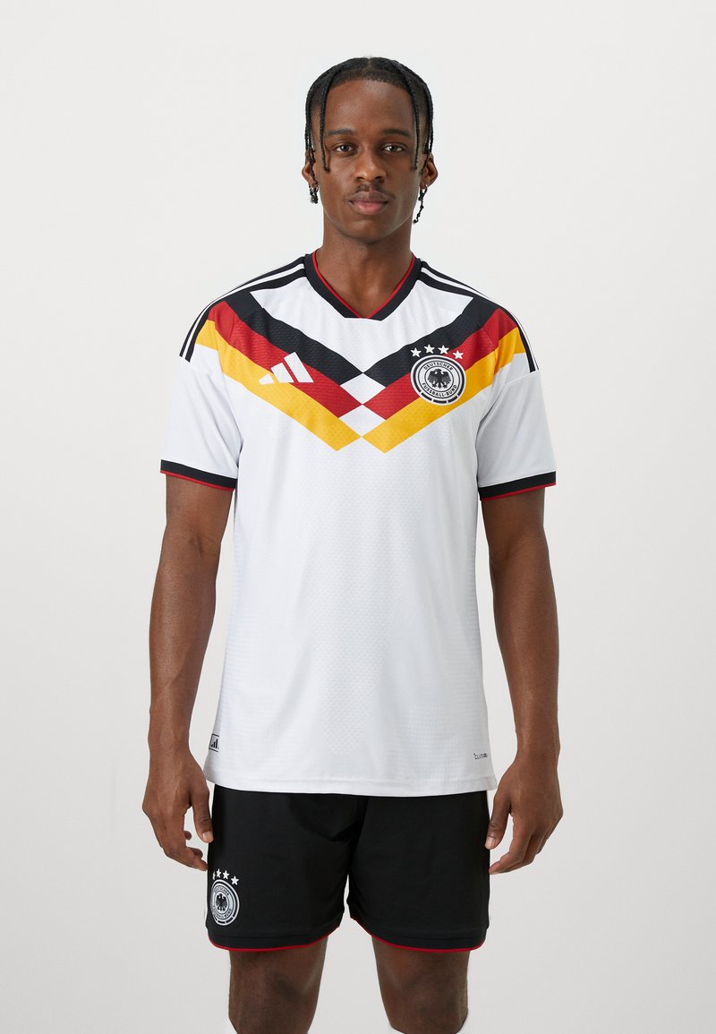 Mann trägt deutsches Fußballtrikot mit weißem Trikot, das schwarze, rote und gelbe Keile sowie schwarze Shorts mit Teamemblem zeigt.